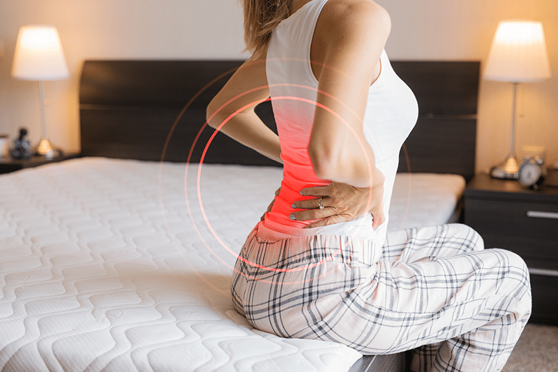 Woman sitting on the edge of a bed holding her lower back in pain due to chronic back pain. | FCE Scan