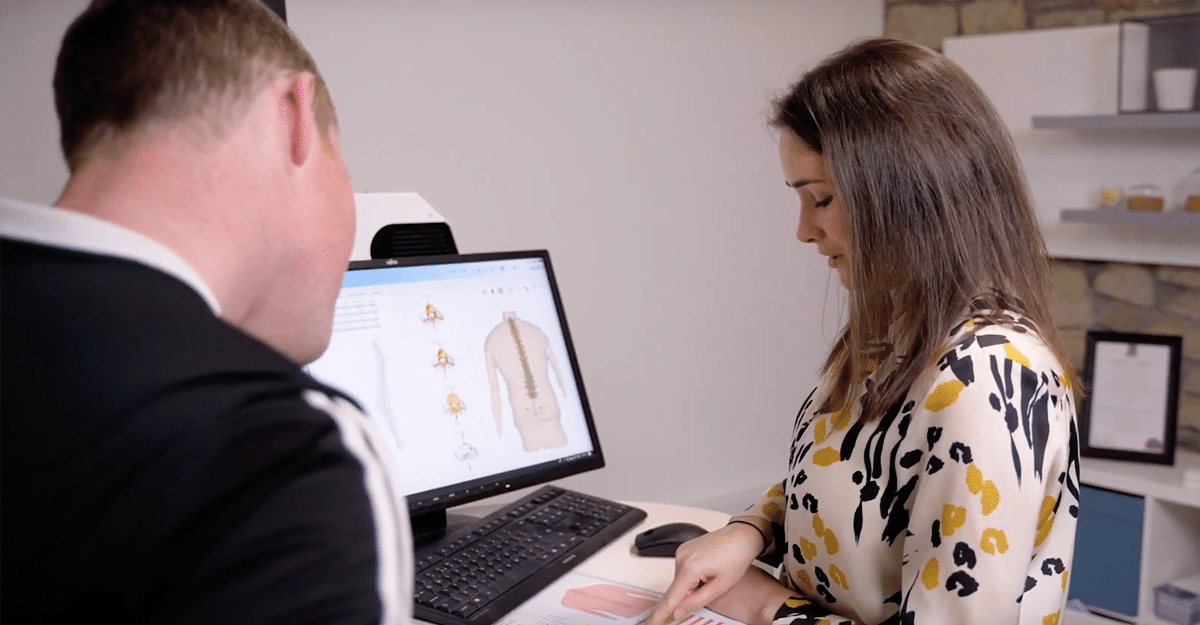 A woman seated at a desk is working on a computer showing graphical content related to an FCE Scan, while a man is seated opposite her with his back facing the camera, appearing to be engaged in | FCE Scan