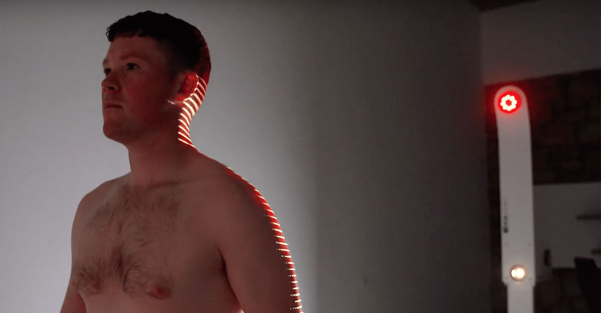 A shirtless man stands indoors with his profile to the camera, looking to the side, illuminated by a red strip of light that casts a defined line along his back during an FCE Scan. | FCE Scan