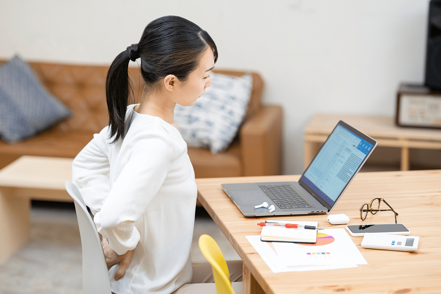 poor-posture A woman sitting at a wooden table, using a laptop and looking over her shoulder with back pain. Papers and stationary are scattered on the table. | FCE Scan