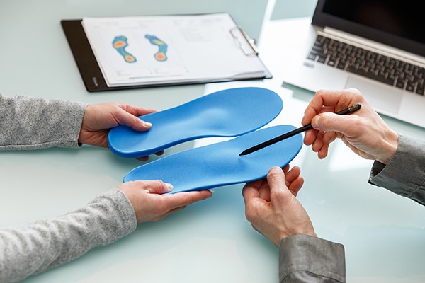 Two people examining blue orthopedic insoles at a desk with a foot diagram and a laptop in the background, deepening their understanding of orthotics. | FCE Scan